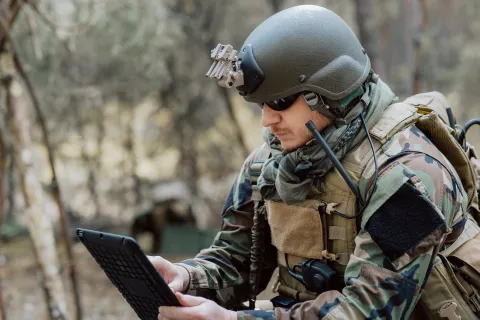 Bearded soldier in uniform sit on military transport crates, analyze data on a tablet and work out tactics at a temporary forest base.