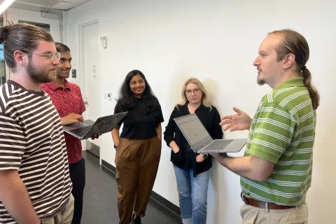 A group of people stand around holding laptops and conversing. 