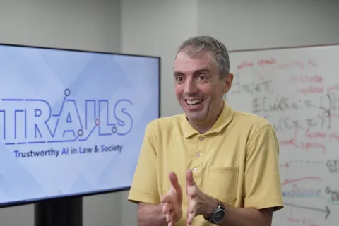 Jordan Boyd-Graber smiles and gestures while seated in front of a screen displaying the logo for "TRAILS: Trustworthy AI in Law & Society" and a whiteboard with handwritten notes.