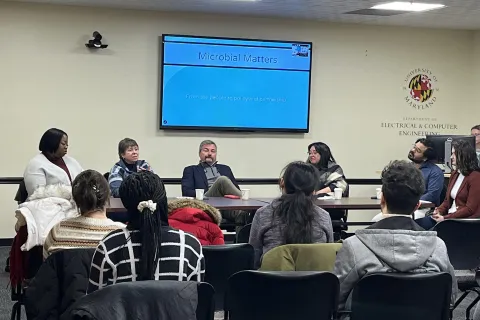 A panel discussion on microbial matters, with several speakers seated at a table, and an audience in chairs facing them. A presentation slide is displayed on the wall behind the speakers.