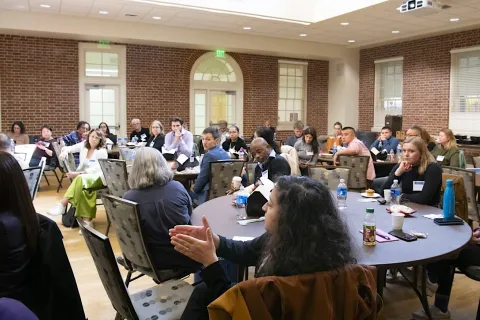 A large group of people seated in a room arranged with tables, some engaged in conversation while others appear to be listening attentively.