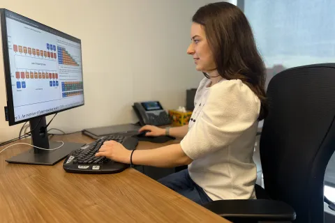 Sarah Wiegreffe sits at a wooden desk, working on a computer with various charts and graphs displayed on the monitor.