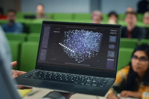 A person holds up a laptop displaying a glowing 3D neural network visualization to an audience of students in a lecture hall.