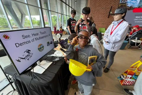 A young boy smiles while trying on smart glasses at a "Demos of Multisensory AI" exhibition booth during Maryland Day 2026.