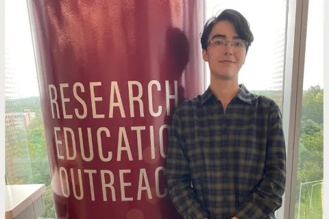 Milijana Surbatovich stands next to a large red pillar featuring the words "RESEARCH EDUCATION OUTREACH" in white text.