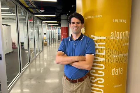 Michael Gullans posing in a modern glass hallway next to a yellow pillar printed with tech terms like "autonomous robotics," "deep learning," and "DISCOVERY."