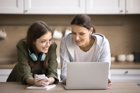 A smiling woman and a teenage girl looking together at an open laptop screen on a kitchen table.