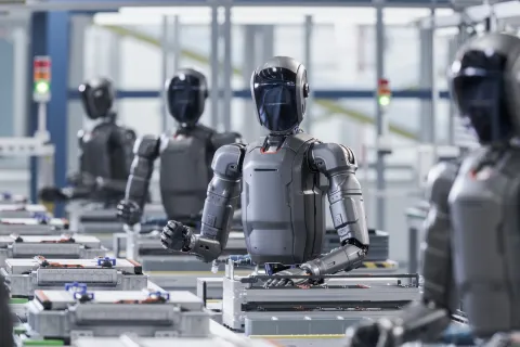 A line of sleek, dark gray humanoid robots working on an assembly line in a modern, high-tech manufacturing factory.