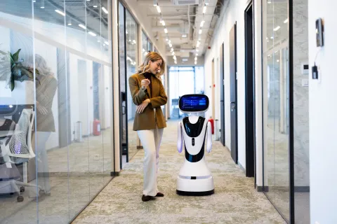 A woman in a tan blazer and light trousers looks down smiling at a modern, white, wheeled service robot standing beside her in a long, glass-walled office corridor. The robot features a lit blue display interface for a head.
