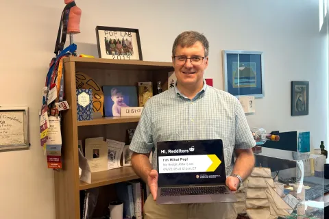 Mihai Pop standing in his office holding an open laptop displaying a Reddit AMA announcement that reads "Hi, Redditors. I'm Mihai Pop!".