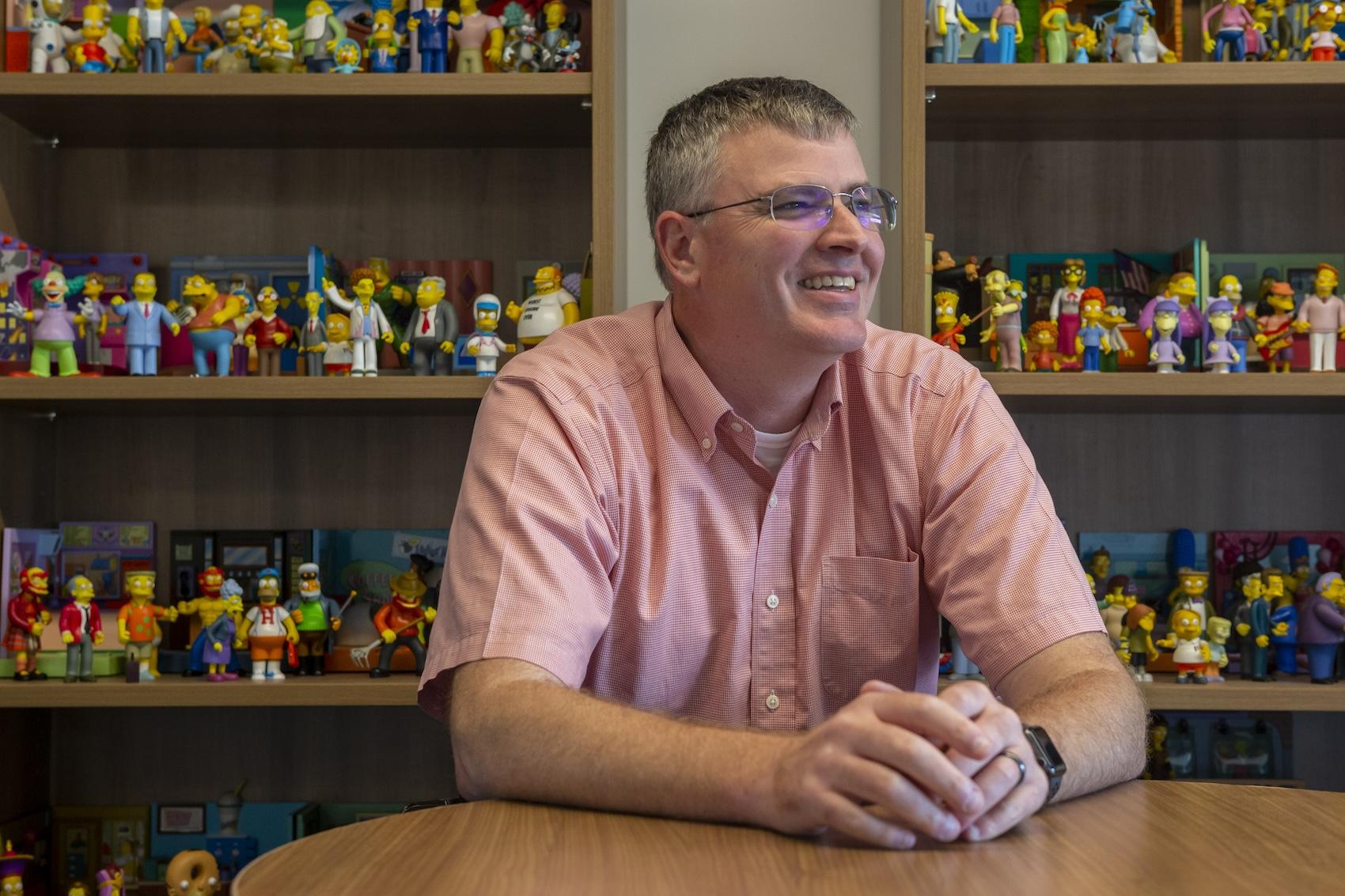 Derek smiling at table with Simpsons figurines in bookshelves behind him
