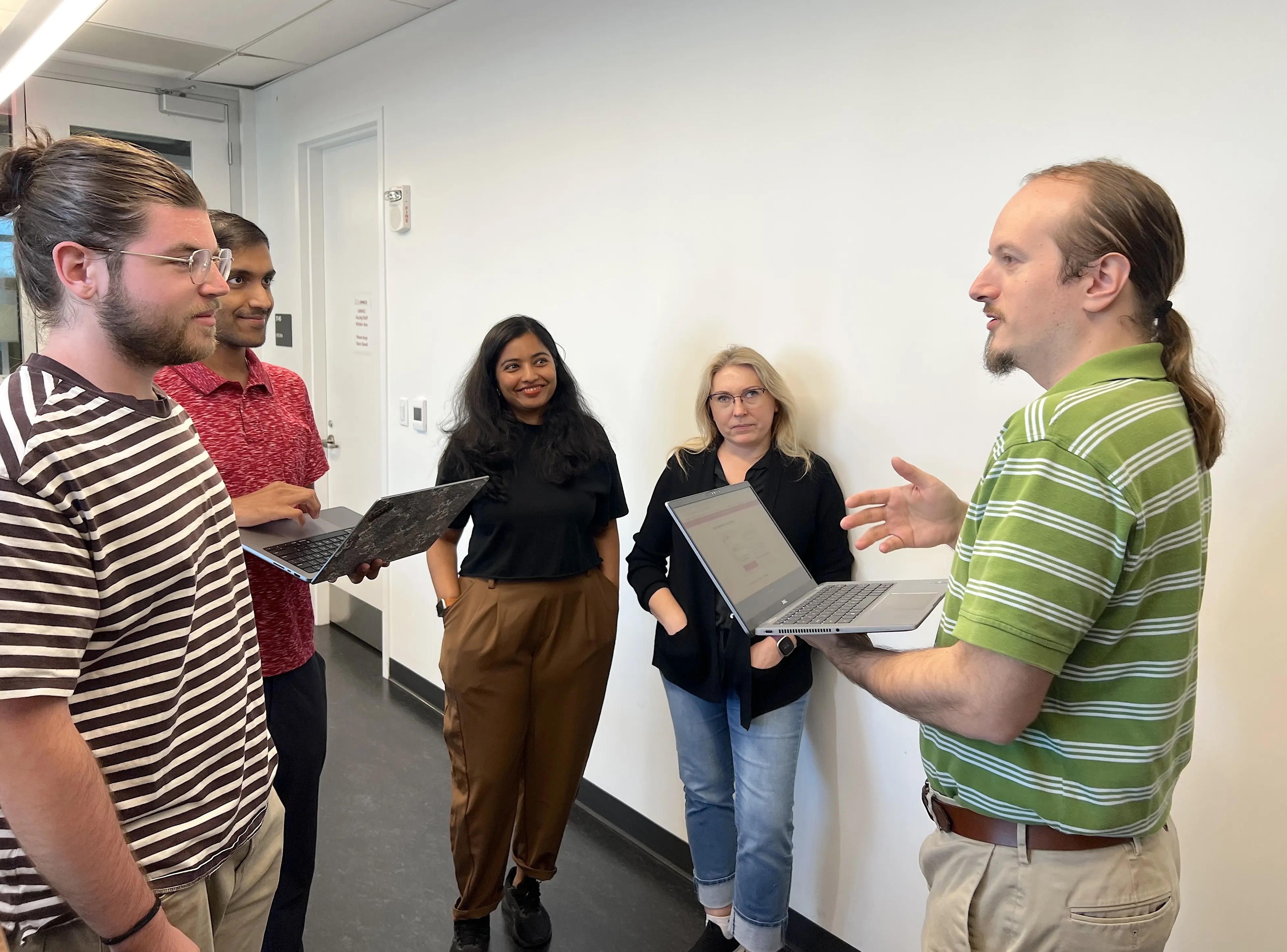 A group of people stand around holding laptops and conversing.