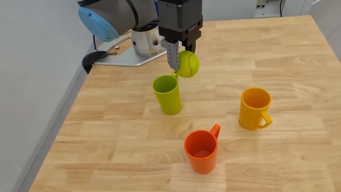 A robotic arm holds a tennis ball on a table surrounded green, orange, and yellow mugs. 