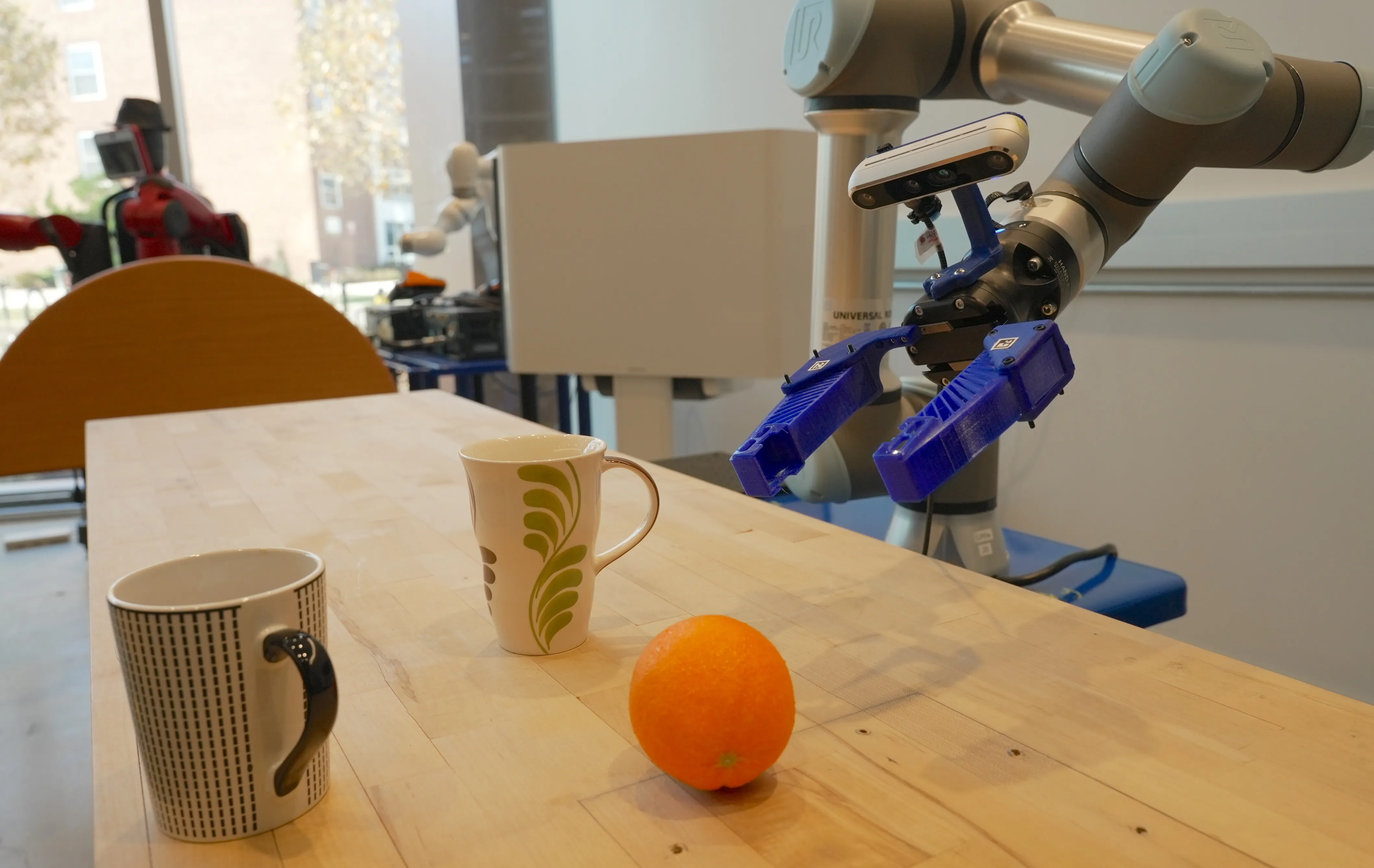 A robotic arm hovers above an orange and two coffee mugs on a wood table. 