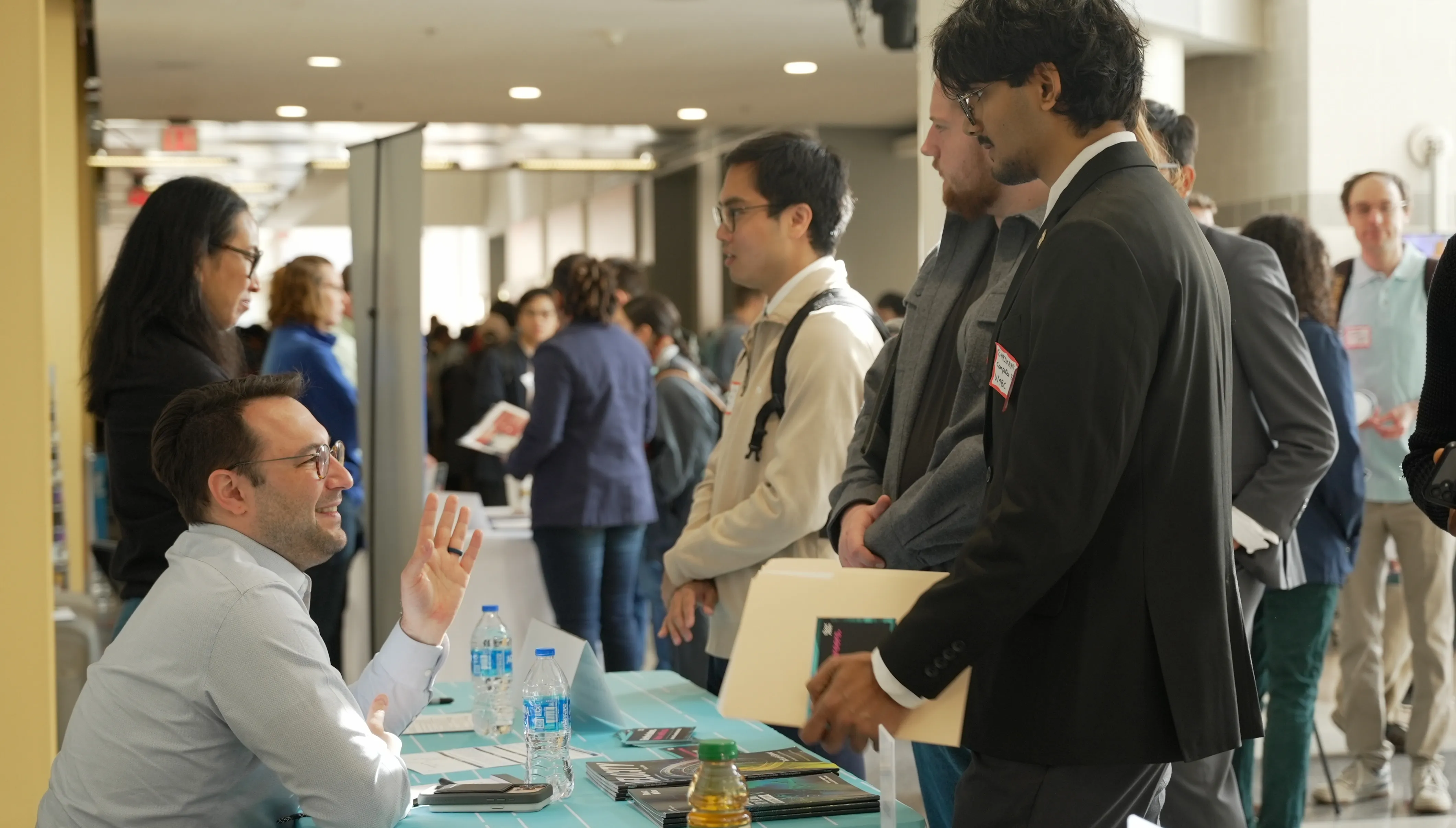 A young man speaks to another man seated at a desk during an event.