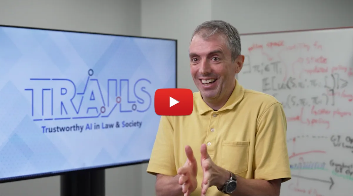 Jordan-Boyd Graber gestures while seated in front of a screen displaying the logo for 'TRAILS: Trustworthy AI in Law & Society' and a whiteboard with handwritten notes.