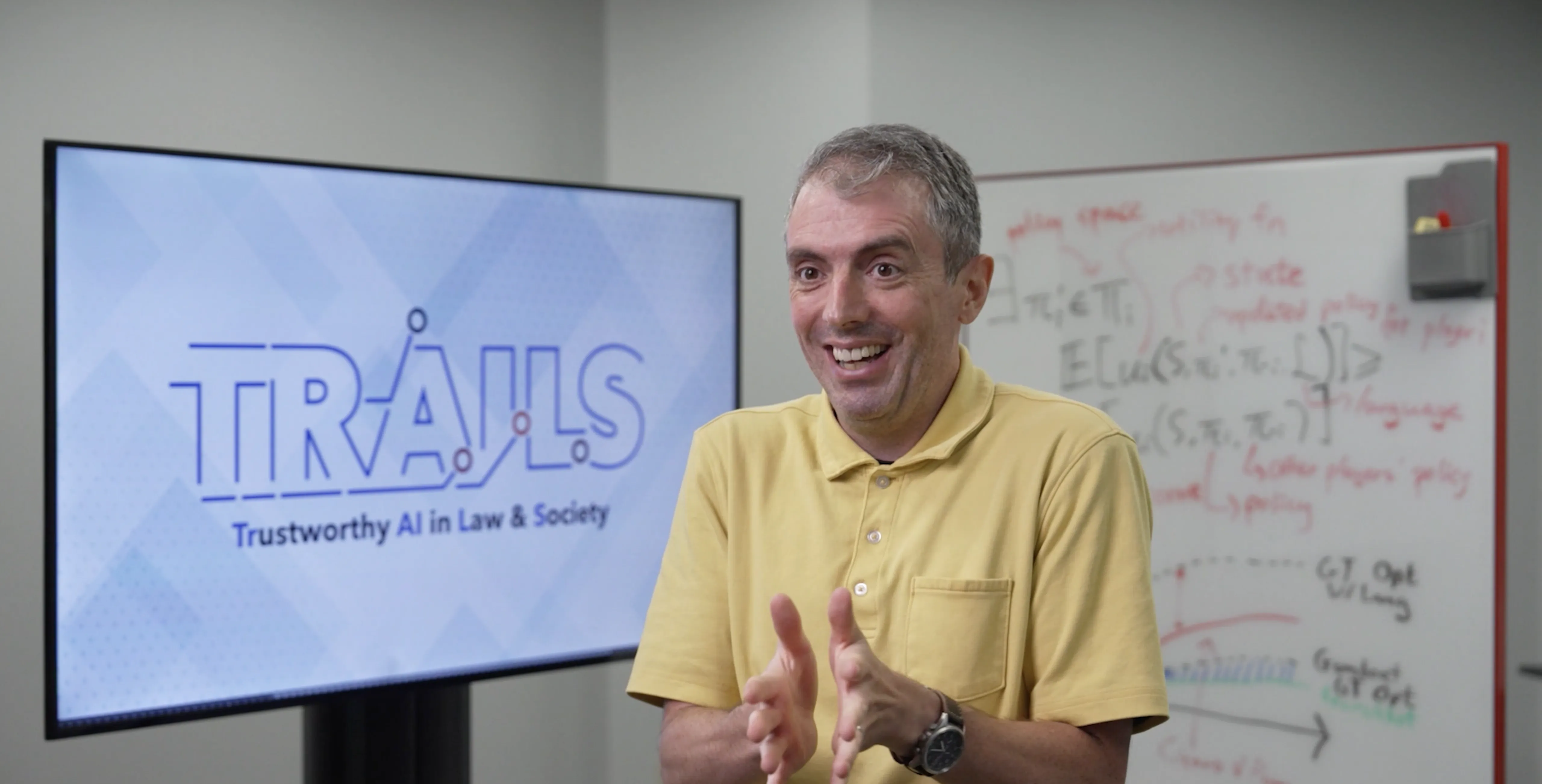 Jordan Boyd-Graber smiles and gestures while seated in front of a screen displaying the logo for "TRAILS: Trustworthy AI in Law & Society" and a whiteboard with handwritten notes.