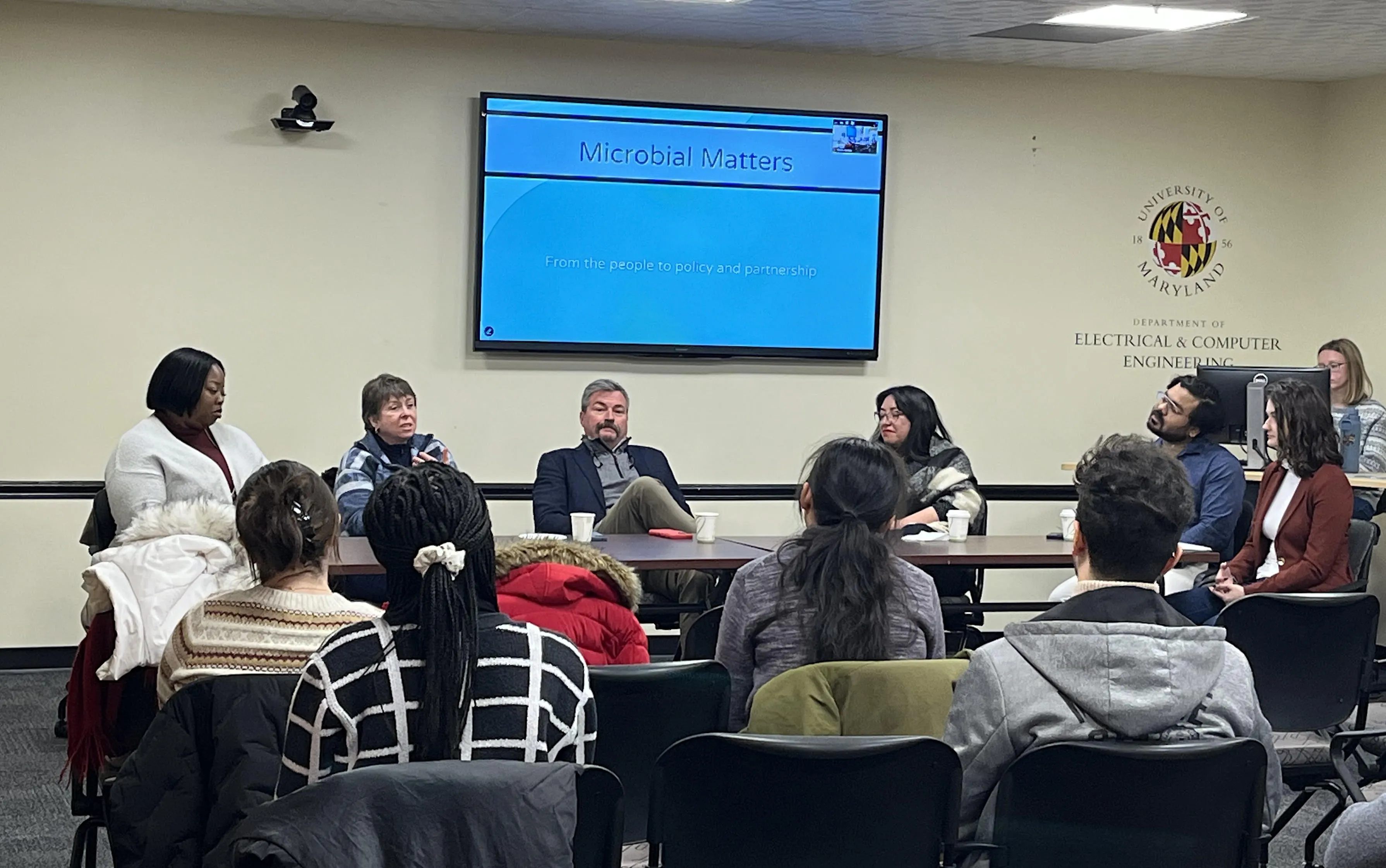 A panel discussion on microbial matters, with several speakers seated at a table, and an audience in chairs facing them. A presentation slide is displayed on the wall behind the speakers.