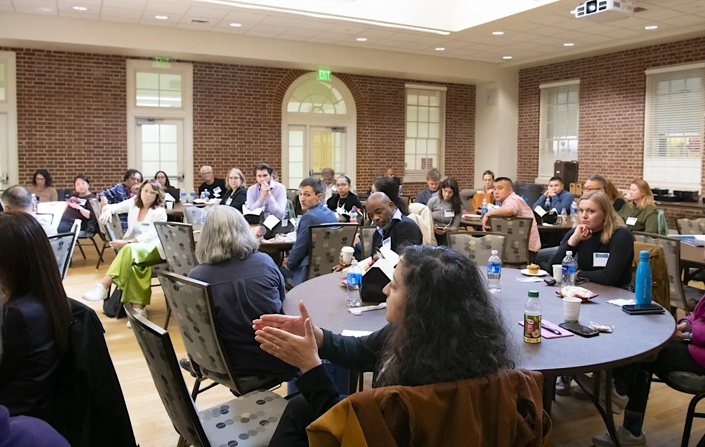 A large group of people seated in a room arranged with tables, some engaged in conversation while others appear to be listening attentively.