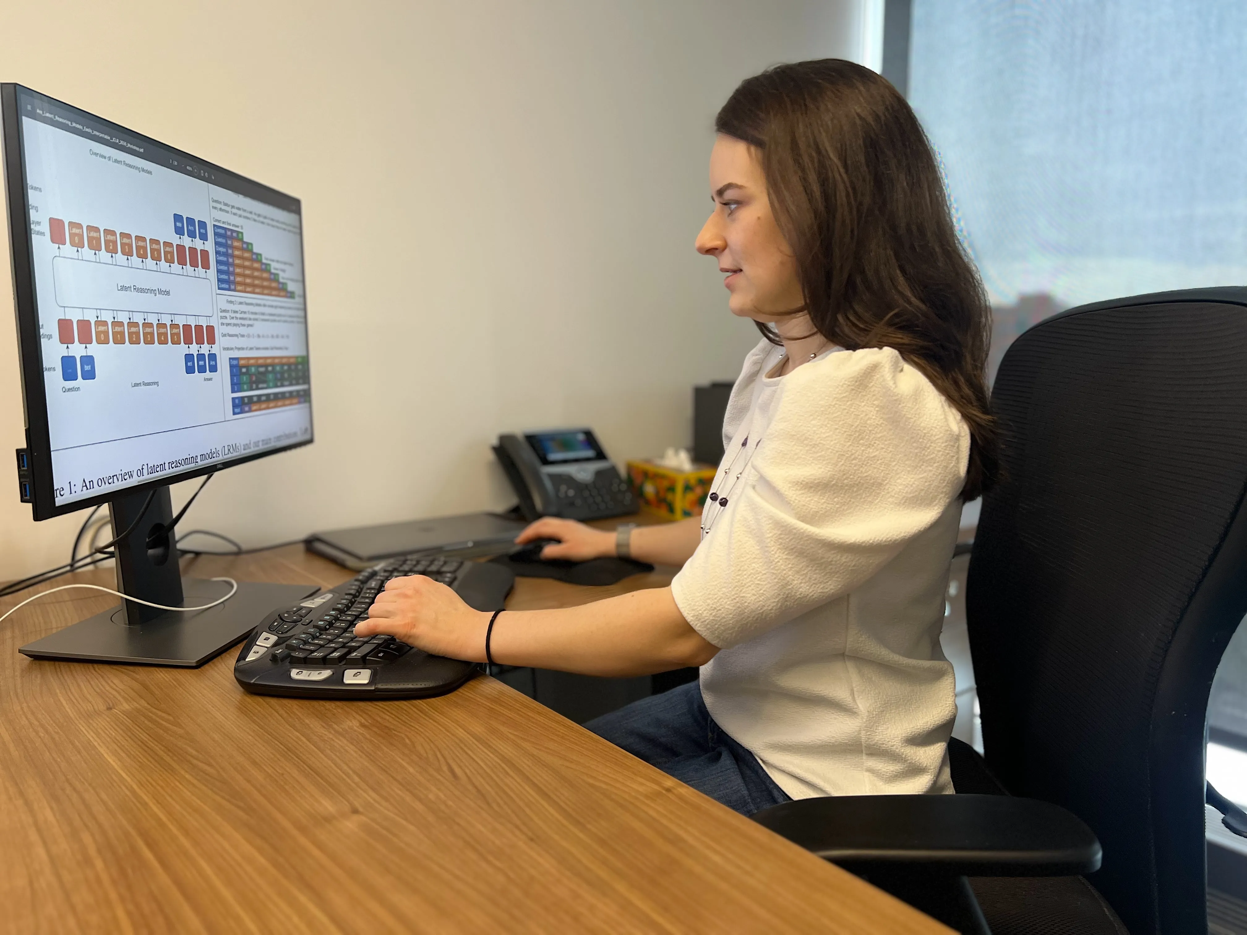 Sarah Wiegreffe sits at a wooden desk, working on a computer with various charts and graphs displayed on the monitor.