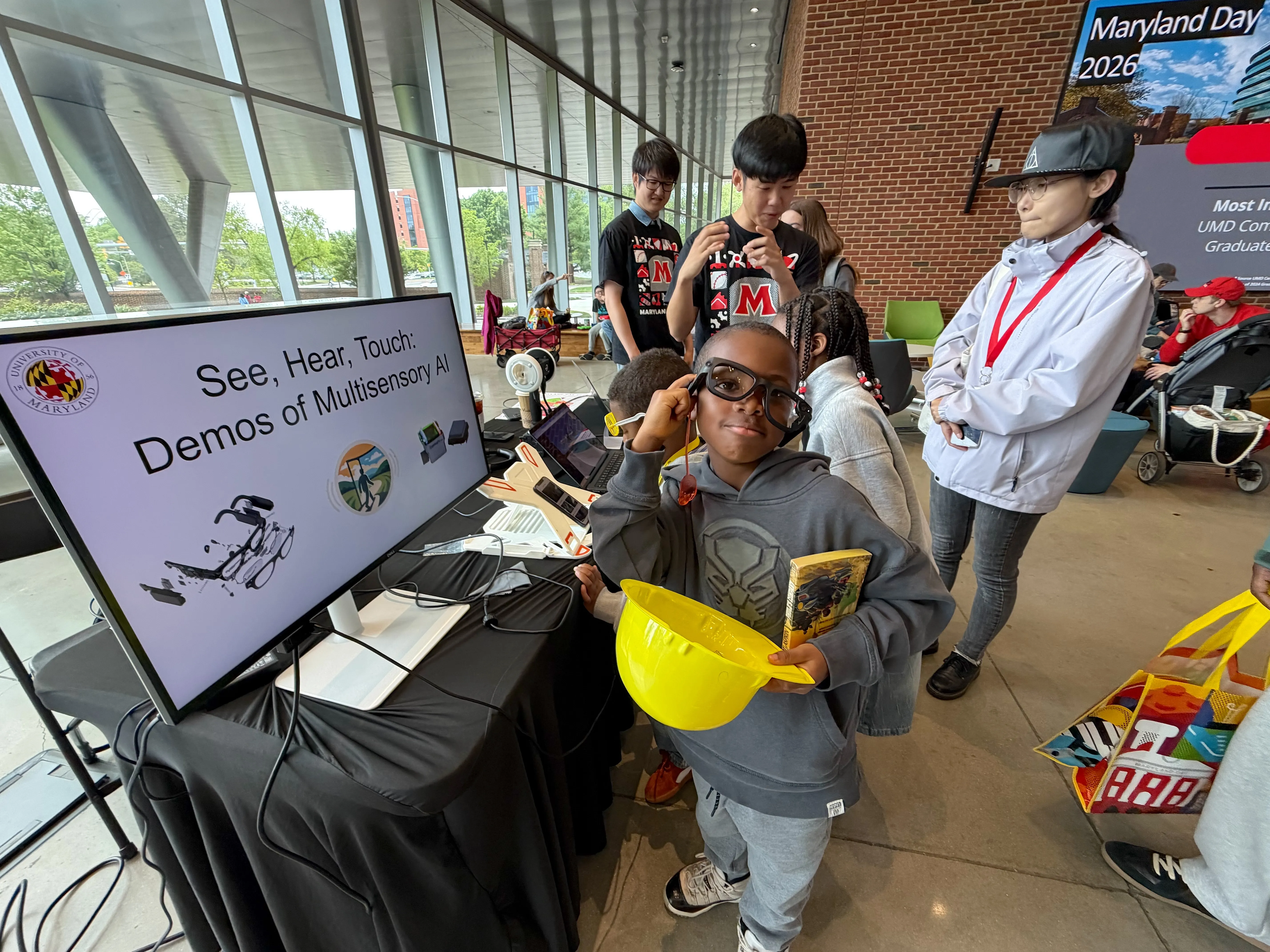 A young boy smiles while trying on smart glasses at a "Demos of Multisensory AI" exhibition booth during Maryland Day 2026.