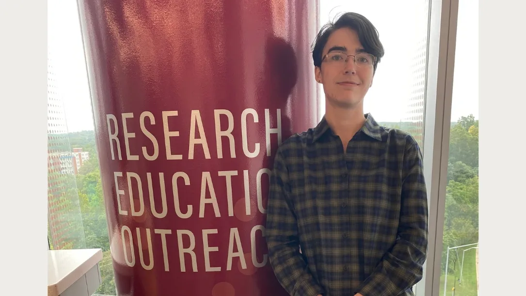 Milijana Surbatovich stands next to a large red pillar featuring the words "RESEARCH EDUCATION OUTREACH" in white text.