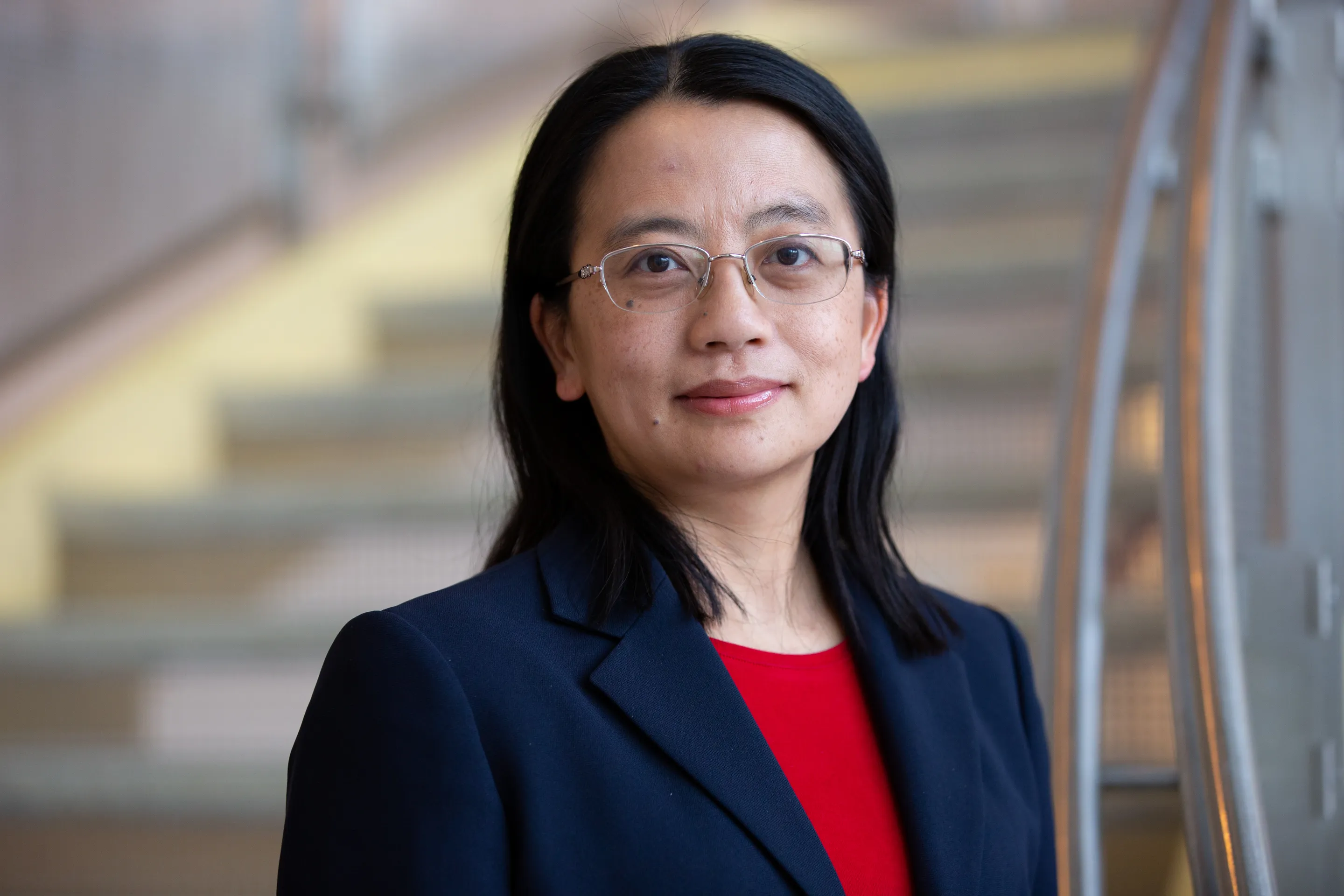 Headshot of Min Wu, a woman with dark hair and glasses, wearing a navy blue blazer over a red top. She is smiling slightly and standing in front of a blurred staircase and metal railing.