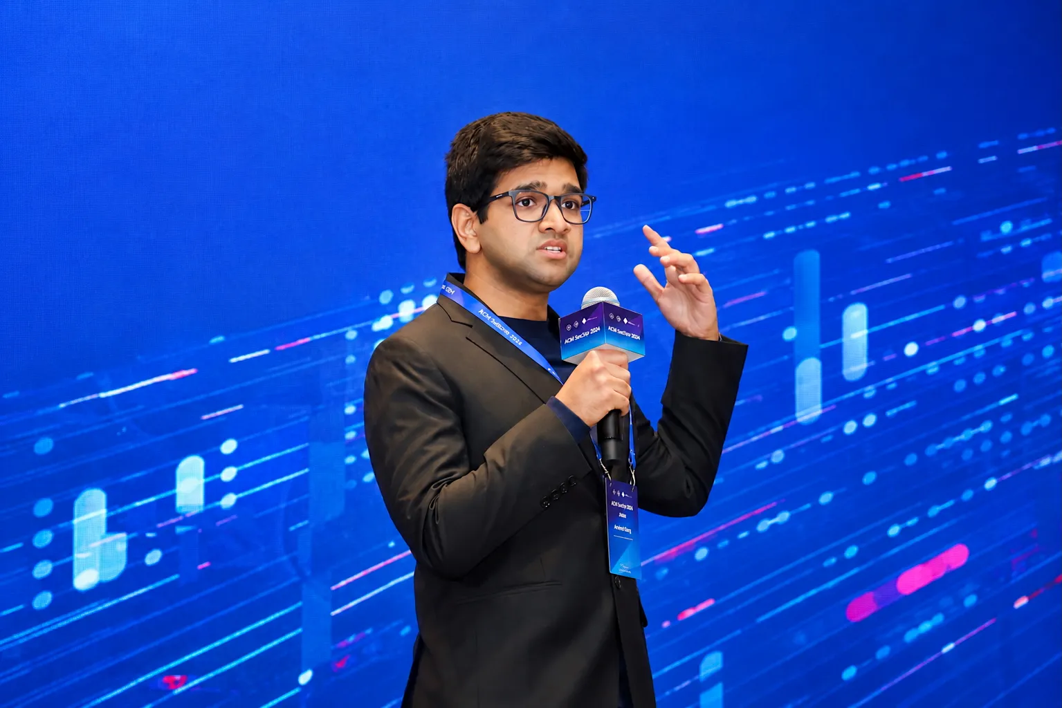 Nakul Garg speaking into a microphone while presenting at an ACM event, wearing a black blazer and glasses against a blue digital-themed background.