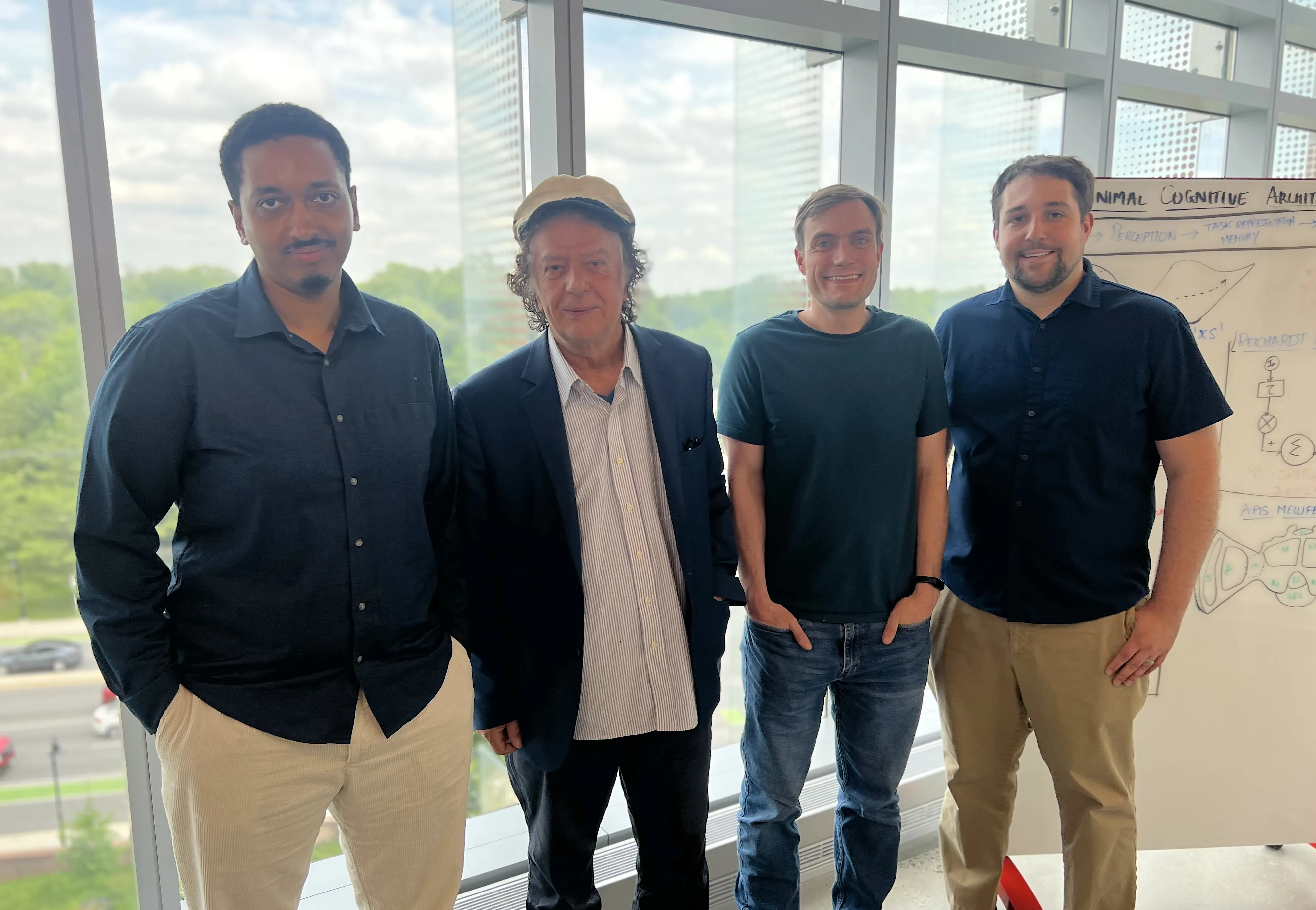 From left to right, Eadom Dessalene, Yiannis Aloimonos, Christopher Metzler, and Levi Burner stand together by a large window for a group photo.