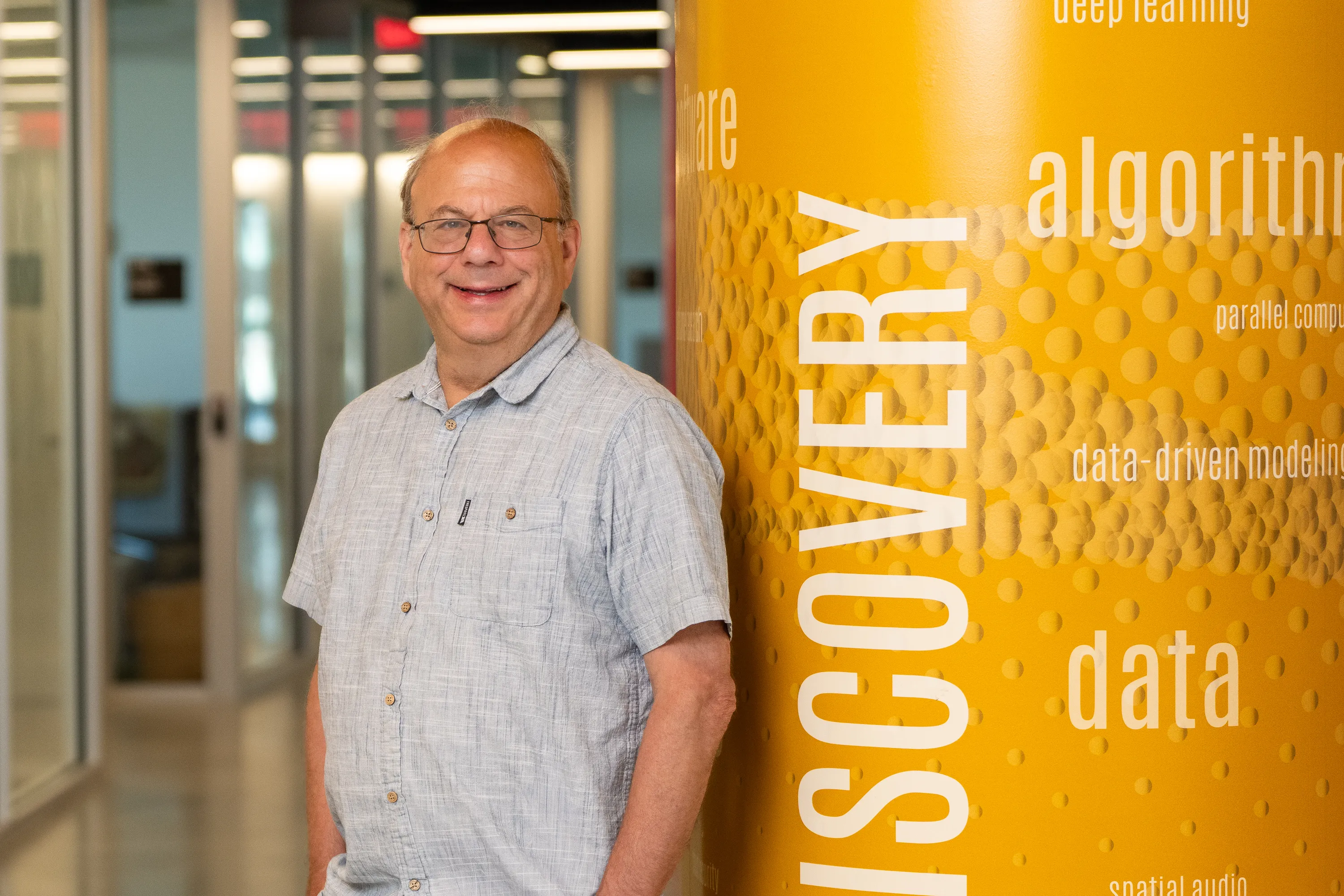 A portrait of David Jacobs, a smiling man with glasses, wearing a light blue short-sleeved button-down shirt. He is standing in a brightly lit office hallway next to a prominent yellow pillar featuring the words "DISCOVERY," "data," and "algorithms."