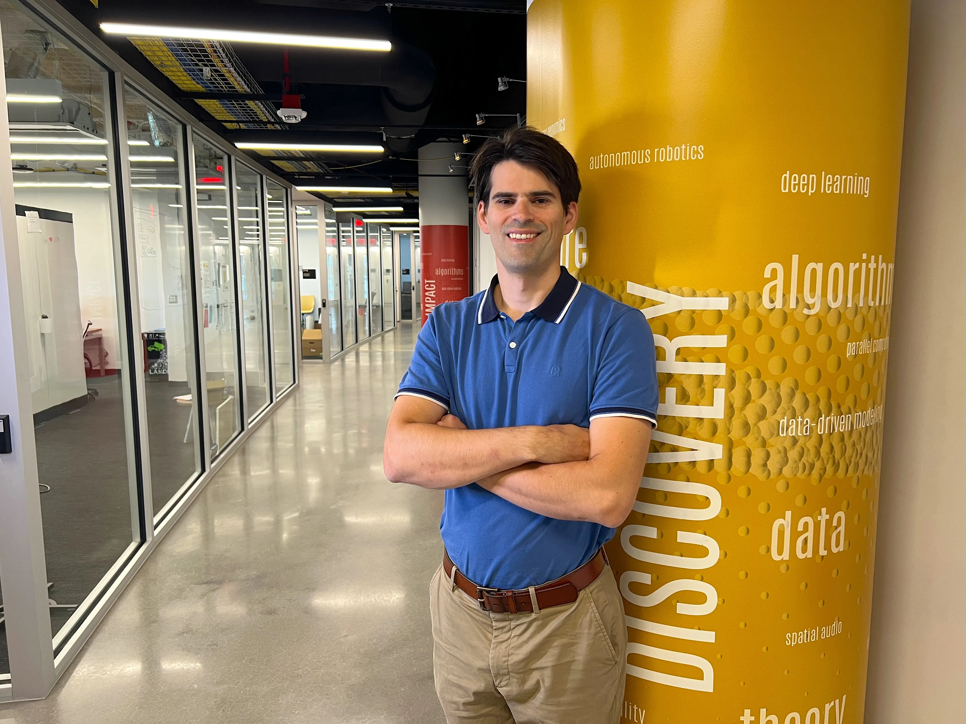 Michael Gullans posing in a modern glass hallway next to a yellow pillar printed with tech terms like "autonomous robotics," "deep learning," and "DISCOVERY."