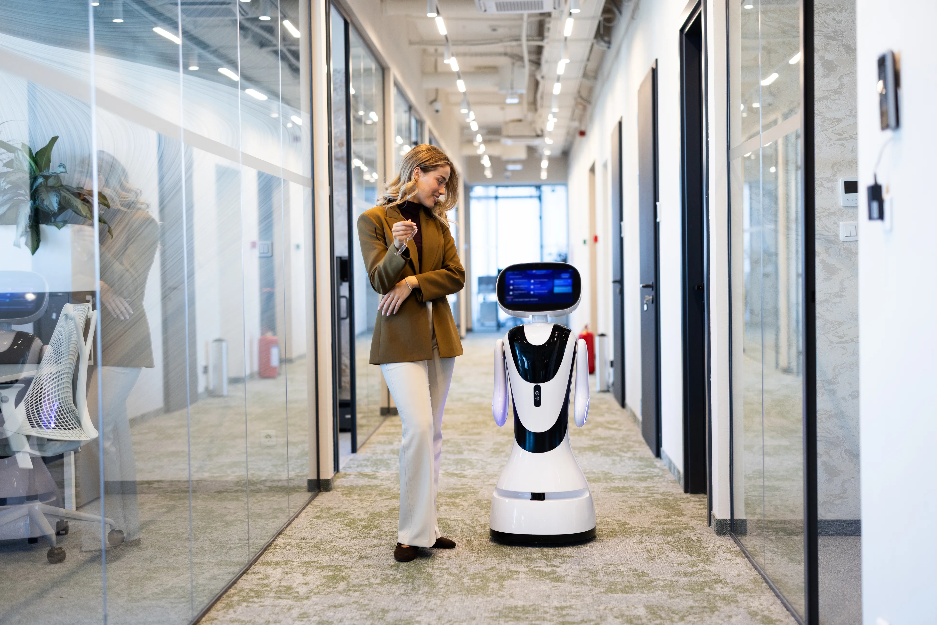 A woman in a tan blazer and light trousers looks down smiling at a modern, white, wheeled service robot standing beside her in a long, glass-walled office corridor. The robot features a lit blue display interface for a head.